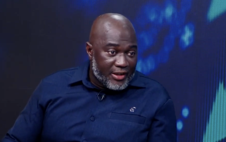African man with a gray beard wearing a dark blue shirt, speaking during a televised interview on a blue-lit set.