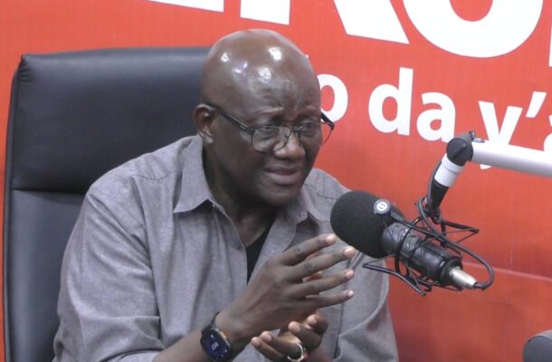 Man wearing glasses and a grey shirt speaks into a large studio microphone, gesturing with his hands in a radio studio backdrop.