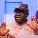 Man wearing a brown cap and glasses speaks while gesturing with both hands during a panel discussion on a colorful studio set.