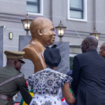Bronze bust statue of a man during a ceremonial unveiling, surrounded by formally dressed attendees outdoors outside a building.