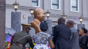 Bronze bust statue of a man during a ceremonial unveiling, surrounded by formally dressed attendees outdoors outside a building.