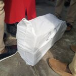 White fabric-wrapped bag on a concrete surface with several people standing nearby, visible legs and shoes around it.