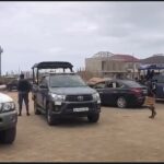 Dusty outdoor parking area with several SUVs and a pickup with a canopy, police vehicles in the background near unfinished buildings.