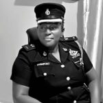 Black-and-white photo of a female police officer in uniform with a name badge reading ‘COMFORT’, seated in a chair.