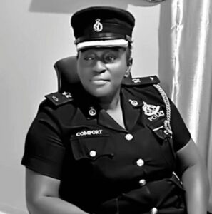 Black-and-white photo of a female police officer in uniform with a name badge reading ‘COMFORT’, seated in a chair.