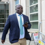 Professional man in navy suit and turquoise tie walking outside a modern building while looking at his phone.
