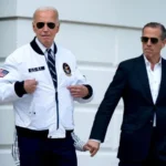Two men wearing sunglasses walk outside a light-colored building; one in a white bomber jacket with patches, the other in a dark suit.