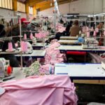 Busy garment factory floor with multiple industrial sewing machines, pink fabric, and workers at their stations.