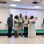 Group of five adults on a stage posing with certificates at an awards ceremony, banner reads NDC in the background.