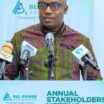 Man in a patterned shirt speaks at a podium with multiple microphones during an Annual Stakeholders event for BUI Power Authority (logo backdrop).