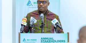 Man in a patterned shirt speaks at a podium with multiple microphones during an Annual Stakeholders event for BUI Power Authority (logo backdrop).
