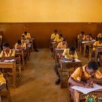 Students in yellow uniforms sit at wooden desks in a classroom, taking a written exam after a test.