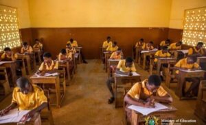 Students in yellow uniforms sit at wooden desks in a classroom, taking a written exam after a test.