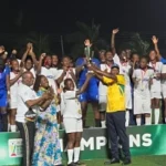 Group of players in white uniforms on a green winners’ podium at night, cheering with raised arms as a presenter in a yellow shirt congratulates them.