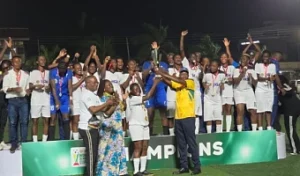Group of players in white uniforms on a green winners’ podium at night, cheering with raised arms as a presenter in a yellow shirt congratulates them.
