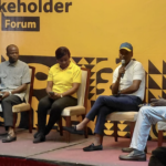 Five panelists sit on a stage during a stakeholder forum; one person speaks into a microphone against a yellow patterned backdrop that says 'Stakeholder Forum'.
