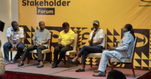 Five panelists sit on a stage during a stakeholder forum; one person speaks into a microphone against a yellow patterned backdrop that says 'Stakeholder Forum'.