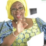 Joyful woman in a yellow headwrap and glasses, gesturing while holding a notebook and speaking.