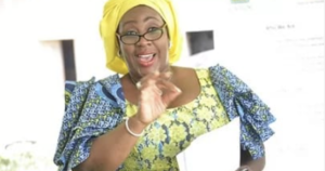 Joyful woman in a yellow headwrap and glasses, gesturing while holding a notebook and speaking.