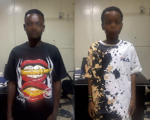 Two young men stand side by side in a hallway; left wears a black shirt with a large open-mouth graphic, right wears a white splatter-pattern FILA shirt.