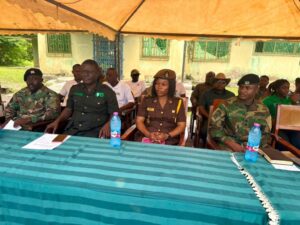 Group of uniformed officials seated under a tent at a long striped table during a formal outdoor ceremony, with water bottles and documents in front of them.