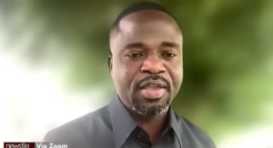 Close-up of a Black man in a dark shirt speaking during a video call, with a blurred green background.