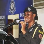 Female police officer in uniform speaks at a podium with a microphone, in front of a blue backdrop and banner.