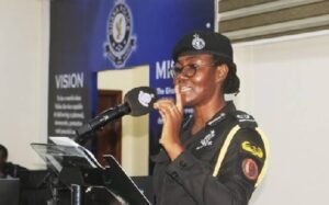 Female police officer in uniform speaks at a podium with a microphone, in front of a blue backdrop and banner.