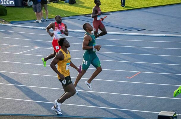 Four male sprinters race on a blue track, wearing colorful uniforms and bib numbers, mid-stride as they surge forward.
