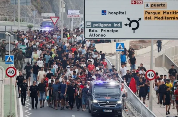 Large crowd of people walking on a road with a police van at the front with flashing lights. Overhead road signs and a 40 km/h speed limit sign are visible.