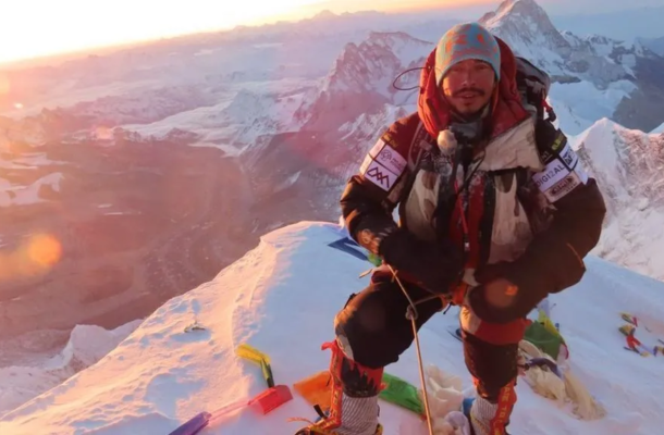 Climber in heavy winter gear stands on a snow ridge at sunrise with distant mountains.