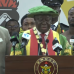 Central figure in a green hat and multicolored ceremonial sash delivering a speech at a KNUST podium, flanked by aides and reporters behind him.
