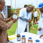 Outdoor demonstration of handwashing products: a man in a brown shirt shows a jar to presenters in white lab coats at a table with several labeled bottles, a microphone nearby.