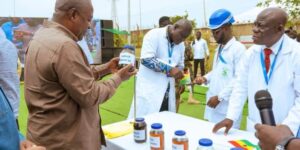 Outdoor demonstration of handwashing products: a man in a brown shirt shows a jar to presenters in white lab coats at a table with several labeled bottles, a microphone nearby.