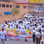 Large crowd of people in white shirts marching down a busy street, holding a colorful banner for a peace walk event.