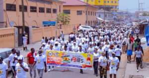 Large crowd of people in white shirts marching down a busy street, holding a colorful banner for a peace walk event.