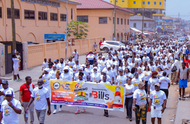 Large crowd of people in white shirts marching down a busy street, holding a colorful banner for a peace walk event.