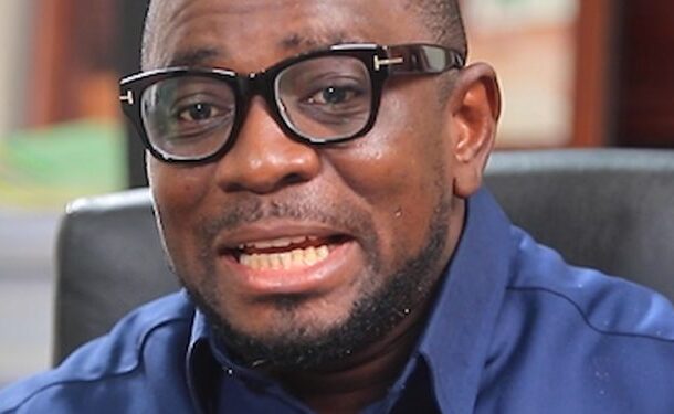 Close-up of a man wearing black-framed glasses and a blue shirt, speaking to someone off-camera.