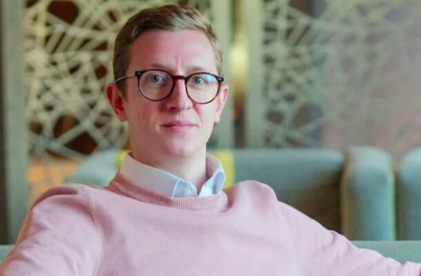 no_translation_7524_e0cy2fufqun7bgm01pwyz9lm_no_translation_translation | Ghanaguardian.com Man with short light hair and round glasses wearing a pink sweater and light blue collared shirt, seated in a modern lounge area.