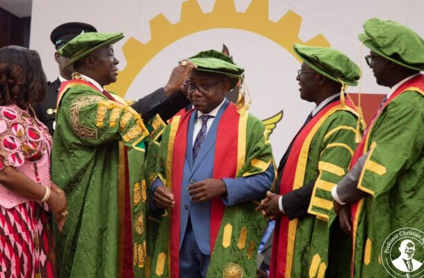 Group of graduates in green robes with gold trim, a man is hooded by a colleague during a ceremonial ceremony, with a woman in pink beside them and a large yellow gear logo in the background.