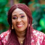 Portrait of a smiling woman with burgundy braids, gold earrings and necklace, wearing a pink floral top outdoors.