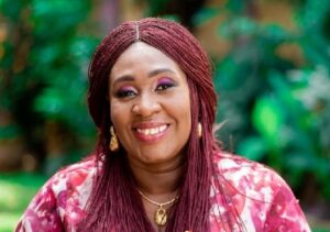 Portrait of a smiling woman with burgundy braids, gold earrings and necklace, wearing a pink floral top outdoors.