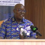 Man in a blue patterned shirt speaks at a podium with multiple microphones during a press conference, with a banner in the background reading University of Ghana and ISSER.