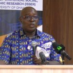 Man in a blue patterned shirt speaks at a wooden podium with multiple microphones, banner behind him indicating ISSER/University of Ghana