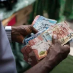 Two hands exchanging a fan of Ghanaian cedi banknotes, including 50‑cedi and smaller denomination bills, outdoors.