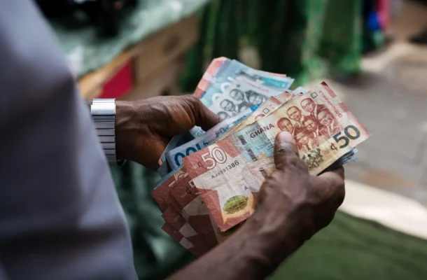 Two hands exchanging a fan of Ghanaian cedi banknotes, including 50‑cedi and smaller denomination bills, outdoors.