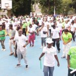 Large crowd participating in an outdoor group exercise on a blue basketball court, mostly in white shirts with some green outfits.