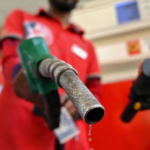 Close-up of a fuel nozzle dripping gasoline, held by a gas station attendant in a red uniform, with a motorcycle in the background.