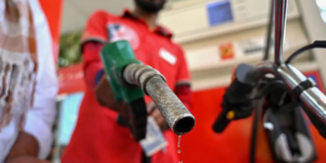 Close-up of a fuel nozzle dripping gasoline, held by a gas station attendant in a red uniform, with a motorcycle in the background.
