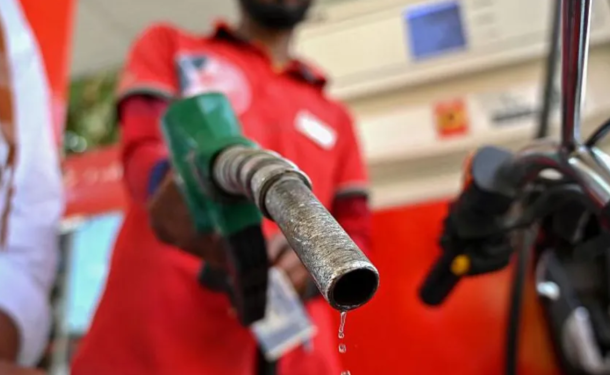 Close-up of a fuel nozzle dripping gasoline, held by a gas station attendant in a red uniform, with a motorcycle in the background.